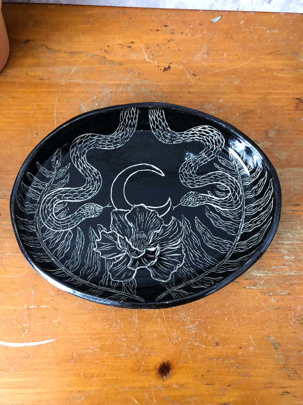It's all about the details with this delightful serpent platter: two snakes sit opposite of each other framing out a night bloom under a crescent moon. Winding up the perimeters of the platter are two ferns.   Hand carved and glazed with a food safe clear so you don't miss a single detail!  Measures approximately 8.5 inches in length and 6.5 inches in width. *Platter warped in the kiln so it does not sit flush and has a slight wobble. For these reasons it has been discounted.